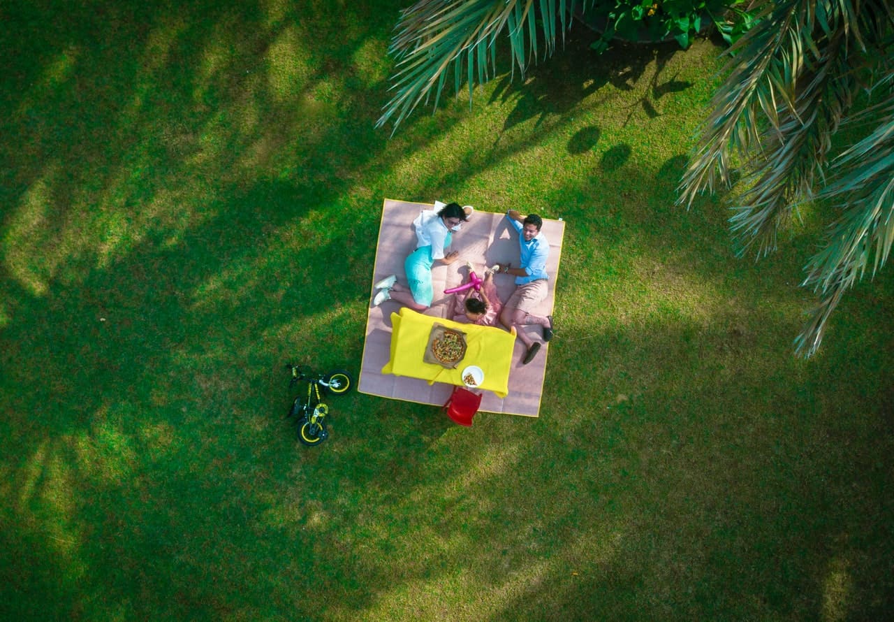 Drone image of a couple with a child laying on a picnic blanket over a beautifully green lawn.